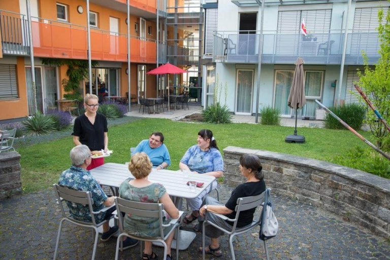 5 Menschen sitzen in einem Garten um einen Tisch. Eine Frau bringt eine Waffel.