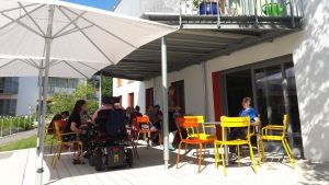 Mehrere Personen sitzen an bunten Tischen und Stühlen unter großen Sonnenschirmen auf einer Terasse.