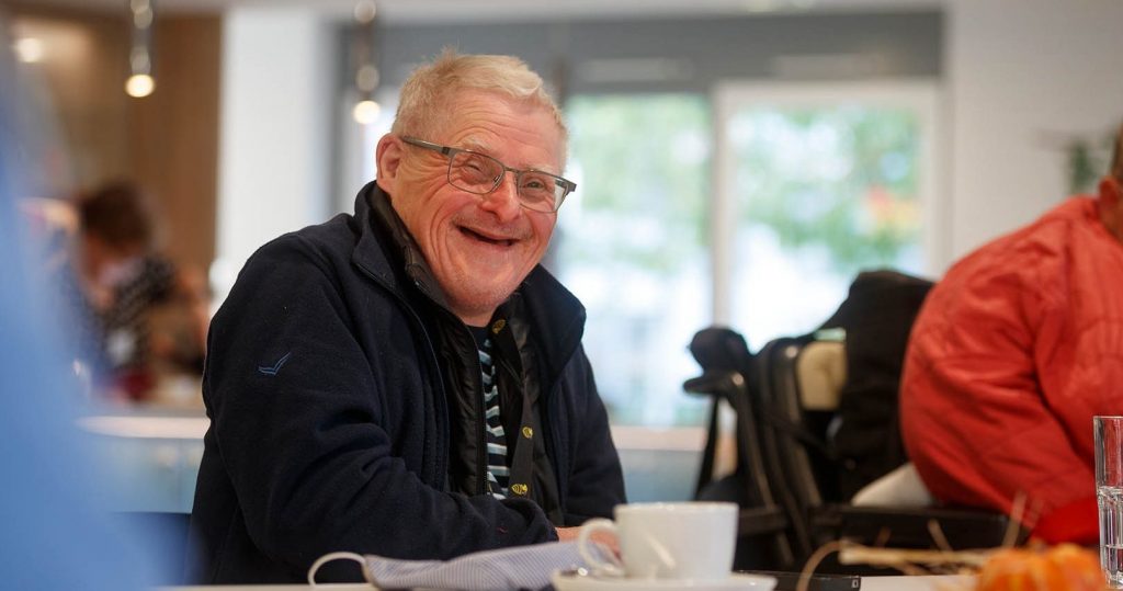 Senior sitzt lächelnd bei einer Tasse Kaffee in heller Seniorentagesbetreuung
