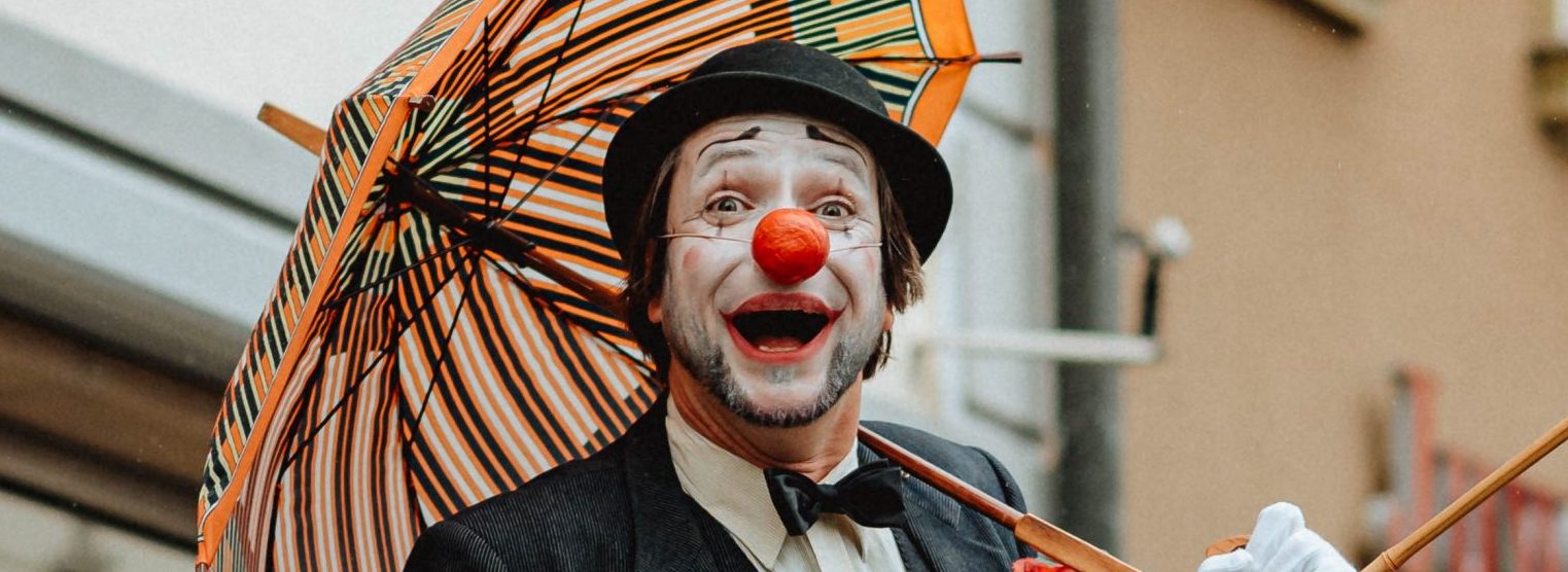 Ein Clown mit einem schwarzen Anzug, weißen Handschuhen, schwarzem Hut und einer roten Nase steht vor Geschäften. IEr hat einen bunt gestreiften Regenschirm aufgespannt.