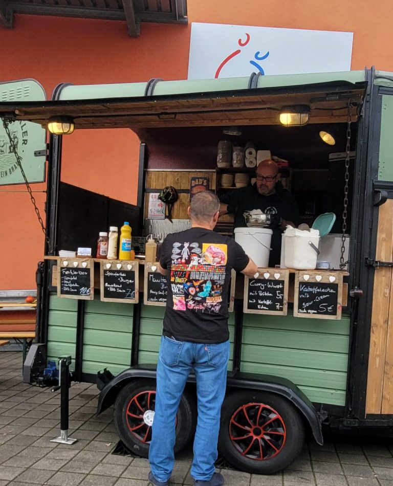 Ein Mann mit einem schwarzen T-Shirt mit buntem Bildern auf dem Rücken steht am Foodtruck.
