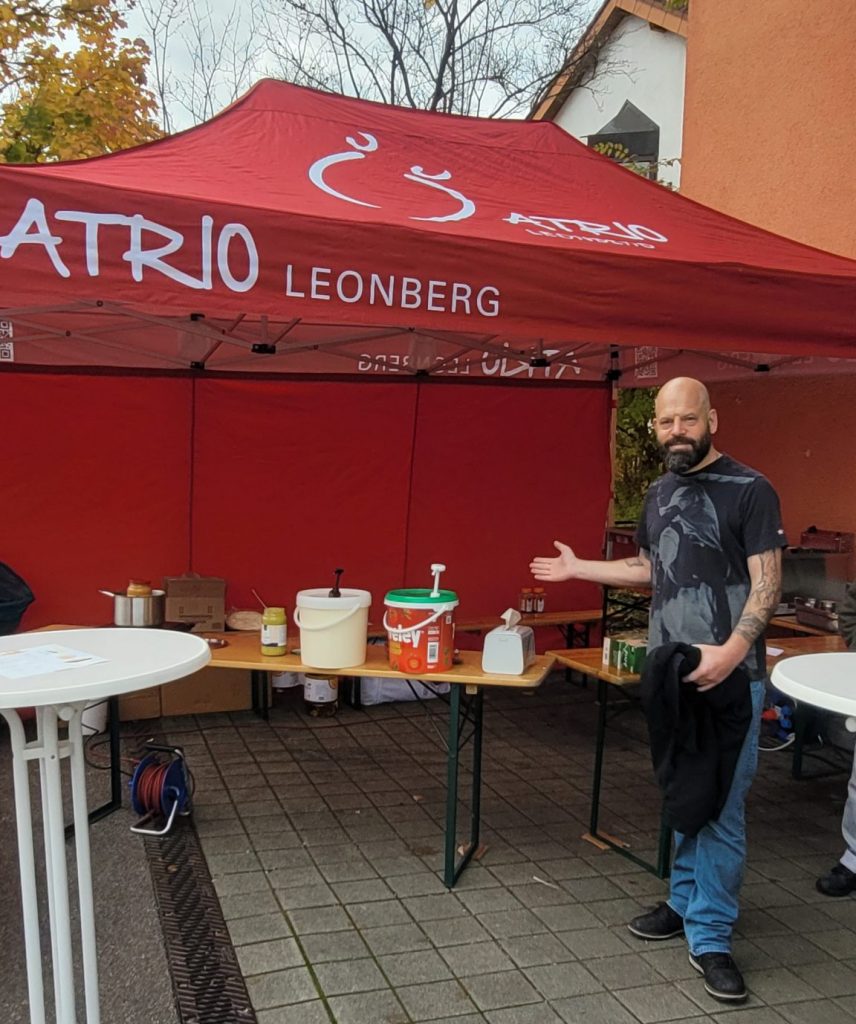 Ein Mann zeigt auf seinen Pommes-Stand unter einem roten Pavillon mit der Aufschrift Atrio.Leonberg.