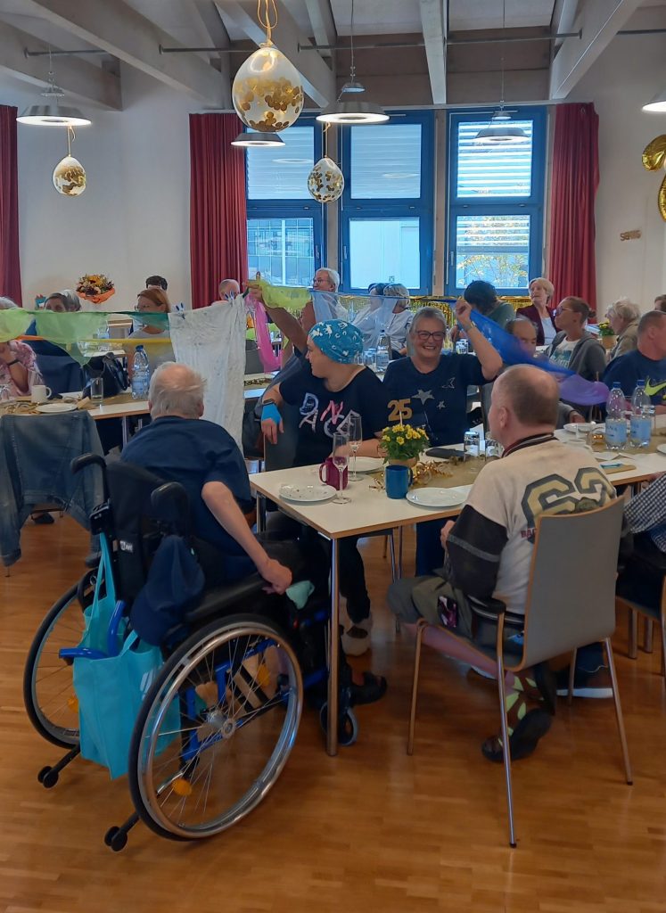 Viele Menschen an bunt gedeckten Tischen. An der Decke hängen silberne Luftballons, in der Mitte des Raums eine Wäscheleine.