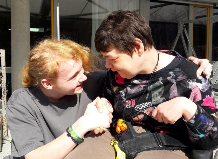 Ein FSJ des FuB und eine Klientin im Rollstuhl lächeln sich an.