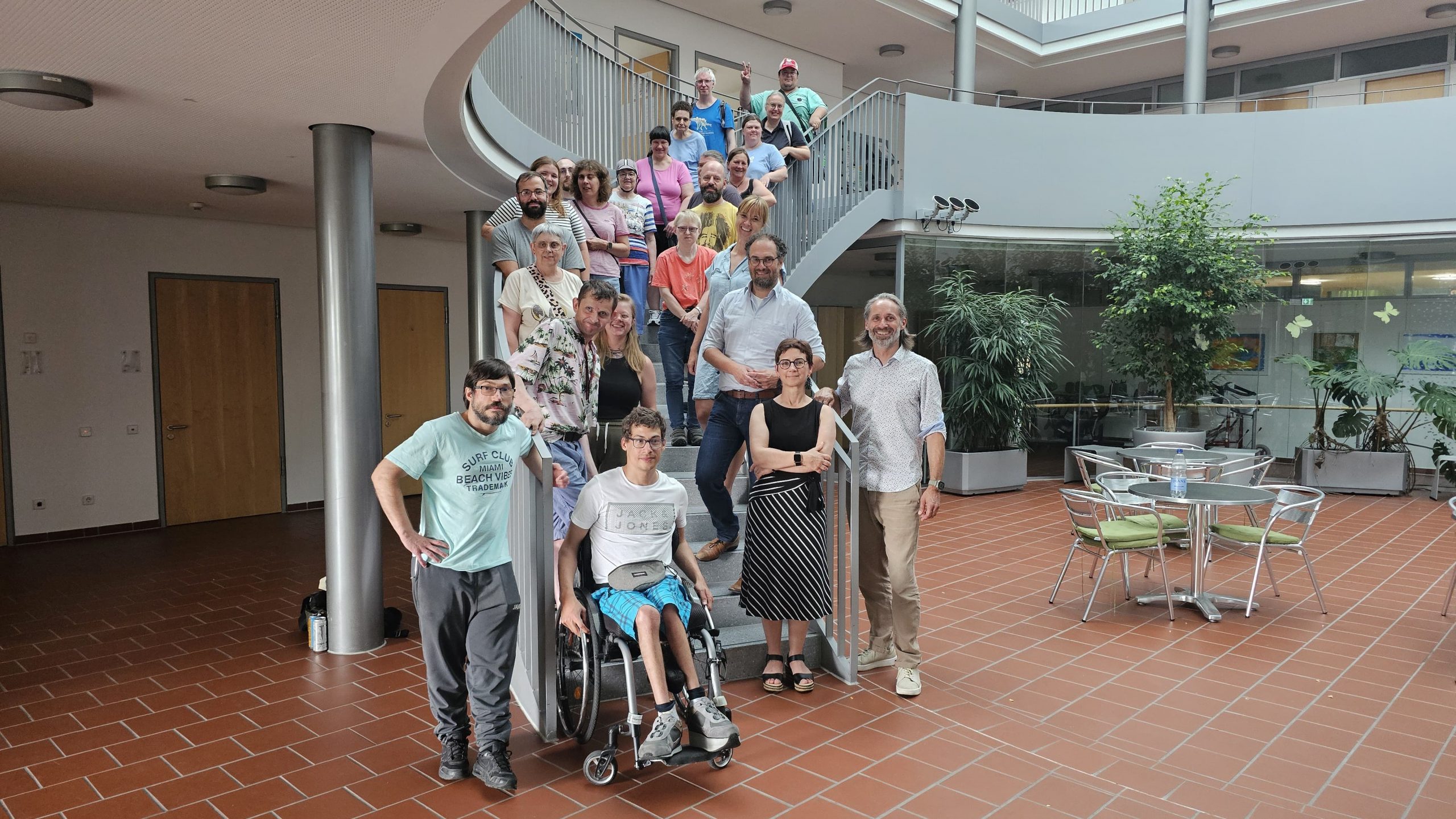 Werkstatträte, Bewohnerbeiräte, Frauenbeauftragte, Vertreter*innen des Aufsichtsrats und Herr Metz stehen auf der Treppe im Haus Atrio.