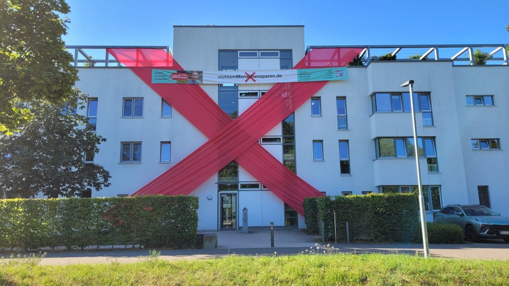 Ein großes rotes Kreuz aus Bändern hängt am Haus Ulmer Straße 58. Darüber der Schriftzug NichtamMenschensparen.de